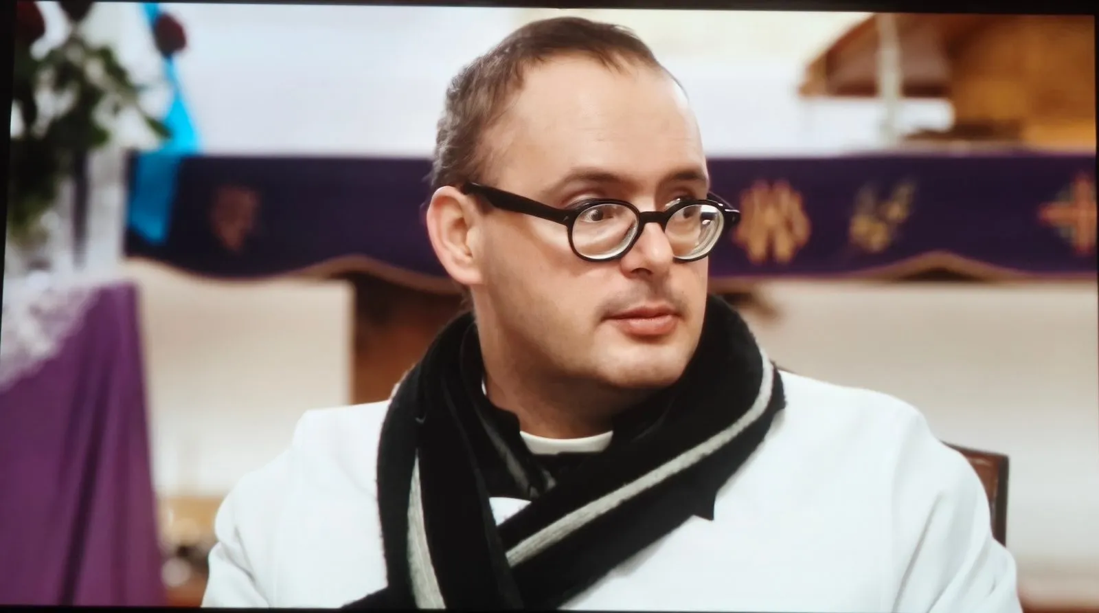 Kadr z filmu: ksiądz Jan Kaczkowski w białej albie i szarym szaliku, w okrągłych okularach, siedzi w kościele przed ołtarzem nakrytym fioletowym suknem