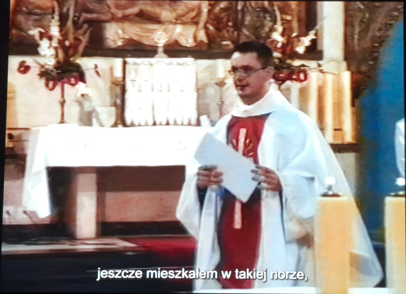 Kadr z filmu: młody ksiądz w białej albie i czerwonym ornacie stoi przy ołtarzu, trzymając w rękach kartkę
