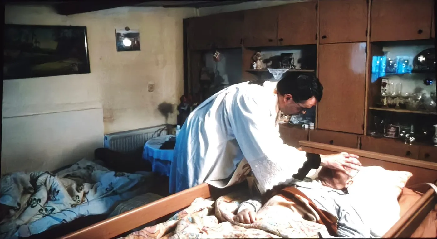 Kadr z filmu: mężczyzna w białej albie pochyla się nad osobą leżącą w łóżku w niewielkim pokoju