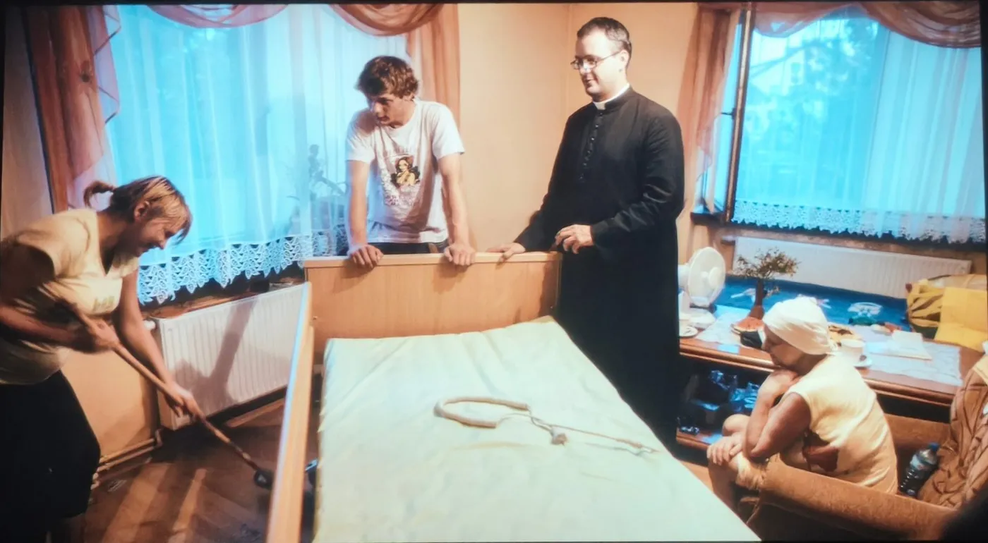 Kadr z filmu: ksiądz w sutannie stoi przy łóżku szpitalnym w mieszkaniu, obok kobieta sprząta podłogę, przy stole siedzi starsza kobieta w chustce